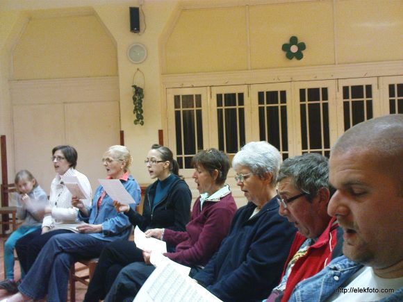 A városi kórus tagjai éppen Daniel Friderici "Jóbarátok" c. négyszólamú művét éneklik a próbán.